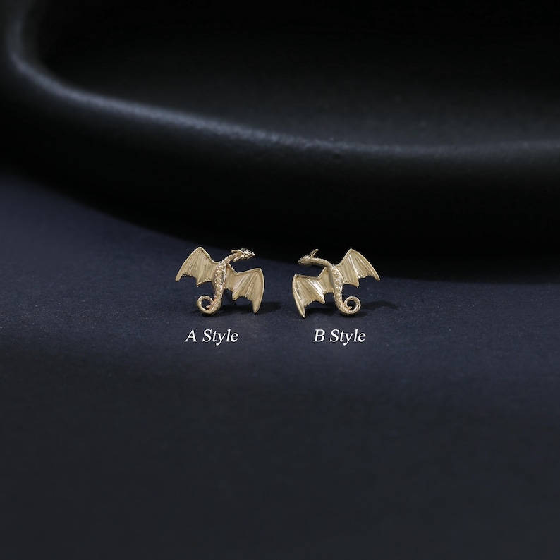 Puede incluir: Dos pendientes de drag&oacute;n dorados, etiquetados como "A Style" y "B Style". Los dragones tienen alas extendidas y detalles. Los pendientes se muestran sobre un fondo oscuro.