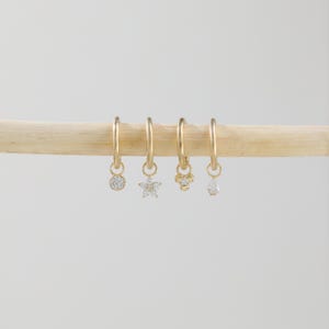 Puede incluir: Cuatro pendientes de aro dorados con diferentes colgantes: un diamante redondo, una estrella, una flor y una l&aacute;grima. Los pendientes cuelgan de una clavija de madera sobre un fondo blanco. Los pendientes son de oro.
