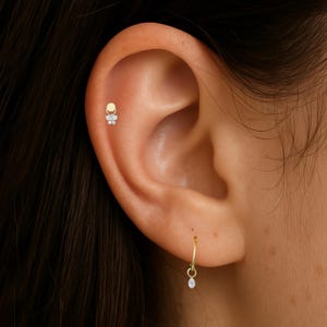 Puede incluir: Primer plano de una oreja adornada con dos joyas. Un peque&ntilde;o pendiente de oro con piedras transparentes est&aacute; en la parte superior de la oreja. Un pendiente de aro dorado con una piedra en forma de l&aacute;grima cuelga del l&oacute;bulo.