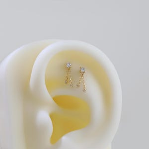 May include: A pair of gold-colored earrings with a dangling design. Each earring features a square-cut, clear gemstone at the top, with a chain of smaller gemstones hanging below. The earrings are displayed against a soft, off-white background.