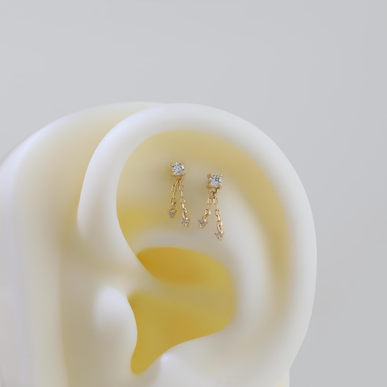 May include: A pair of gold-colored earrings with a dangling design. Each earring features a square-cut, clear gemstone at the top, with a chain of smaller gemstones hanging below. The earrings are displayed against a soft, off-white background.