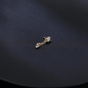May include: A gold-colored earring with a star-shaped top and a dangling chain. The chain holds three small, round, clear gemstones. The earring is set against a dark, textured background.