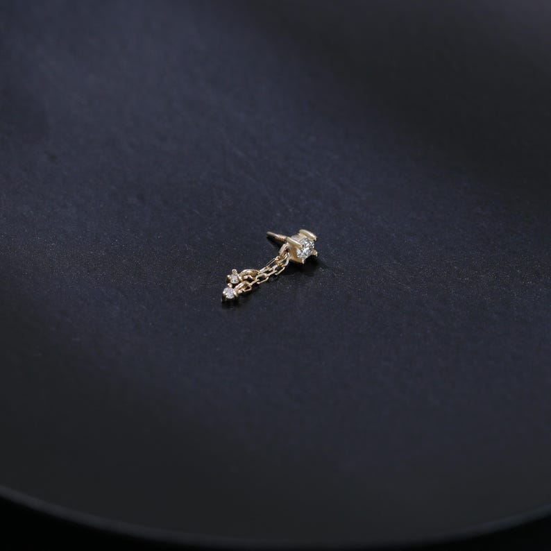 May include: A gold-colored earring with a star-shaped top and a dangling chain. The chain holds three small, round, clear gemstones. The earring is set against a dark, textured background.