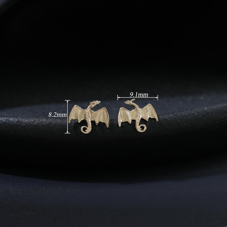 Puede incluir: Dos pendientes dorados con forma de drag&oacute;n. Cada pendiente presenta un dise&ntilde;o detallado de drag&oacute;n con alas extendidas. Los pendientes miden 0,82 cm y 0,91 cm de largo. El fondo es de color oscuro y liso.