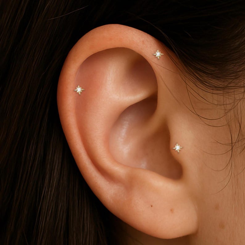 May include: Close-up of a person's ear with three small star-shaped earrings. The earrings are made of gold and have a single clear diamond in the center of each star.