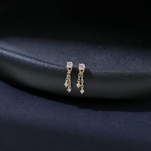 May include: A pair of gold-toned earrings with a square-cut, clear gemstone at the top. Delicate chains dangle from the square, each holding small, round gemstones. The earrings are set against a dark, textured background.