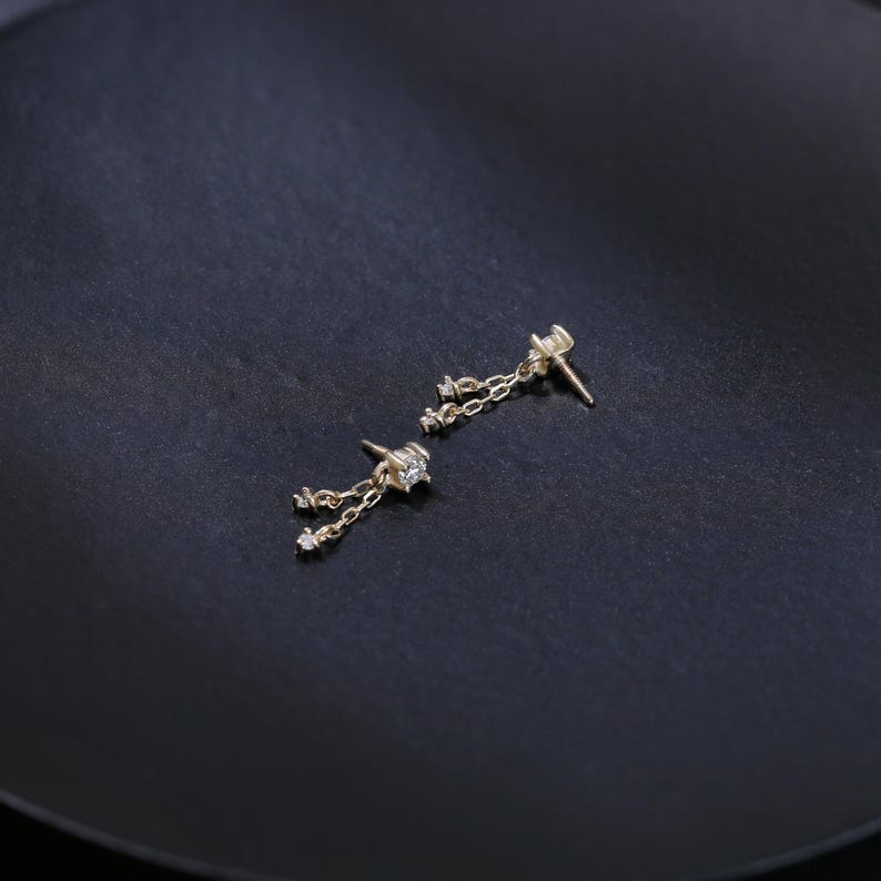 May include: A pair of gold-colored dangle earrings. Each earring features a small, sparkling gemstone at the stud, with a delicate chain suspending smaller gemstones. The earrings are displayed on a dark, textured surface.