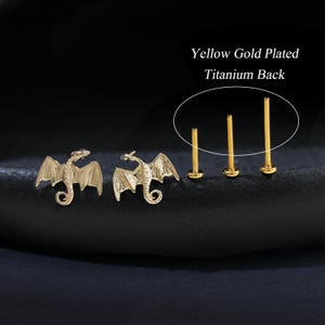 Puede incluir: Pendientes de drag&oacute;n chapados en oro y tres pendientes de bot&oacute;n dorados con parte trasera de titanio. Los pendientes de drag&oacute;n presentan alas detalladas y una cola enroscada. El texto de la imagen dice "Yellow Gold Plated Titanium Back."