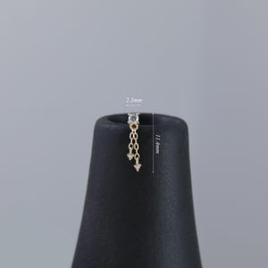 May include: A gold-colored earring with a dangling chain design. The earring features a small, square-cut, clear gemstone at the top, with a chain of small links and two dangling, clear, triangular-shaped gemstones. The earring is 11.4mm long.