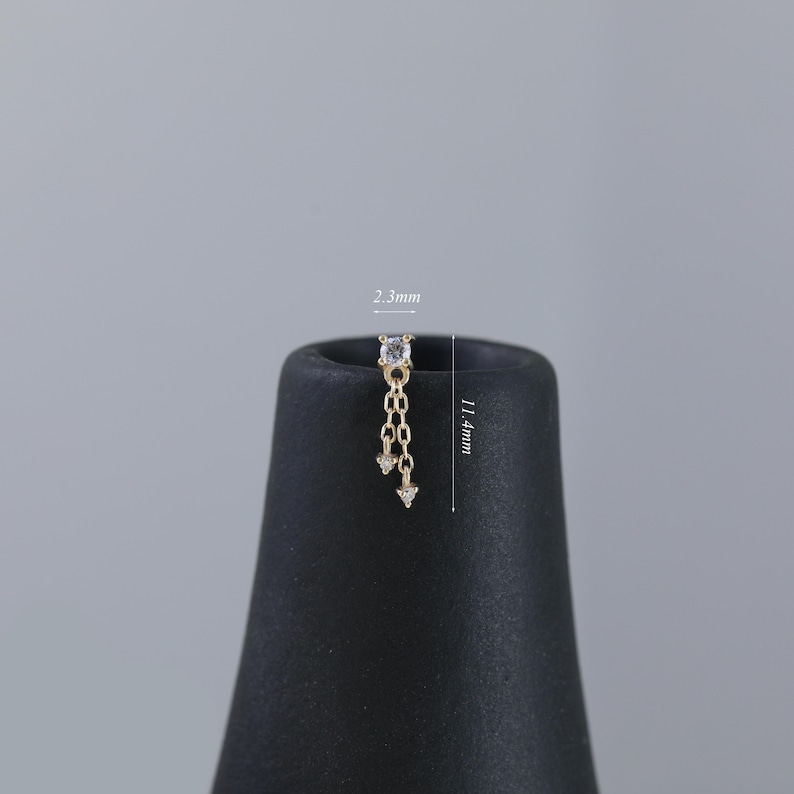 May include: A gold-colored earring with a dangling chain design. The earring features a small, square-cut, clear gemstone at the top, with a chain of small links and two dangling, clear, triangular-shaped gemstones. The earring is 11.4mm long.