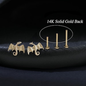 Puede incluir: Pendientes de drag&oacute;n dorados y tres postes de pendientes dorados. Los pendientes de drag&oacute;n tienen alas detalladas y un cuerpo curvado. El texto "14K Solid Gold Back" es visible en la imagen.