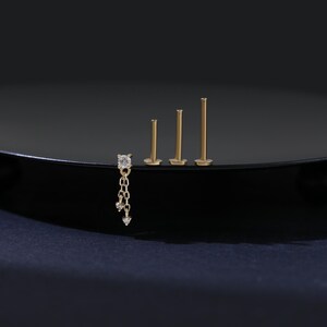 May include: A collection of gold-colored earrings. One earring features a square-cut clear stone with a dangling chain of smaller stones. Three additional earrings are simple posts of varying lengths. The earrings are displayed on a black surface.