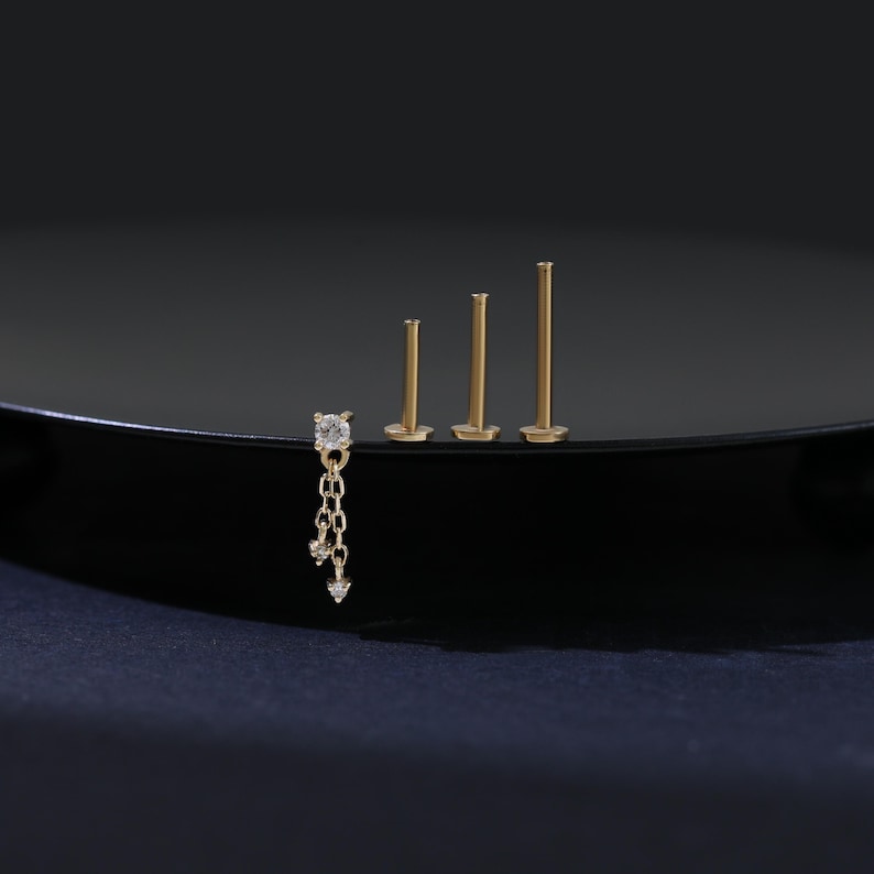 May include: A collection of gold-colored earrings. One earring features a square-cut clear stone with a dangling chain of smaller stones. Three additional earrings are simple posts of varying lengths. The earrings are displayed on a black surface.