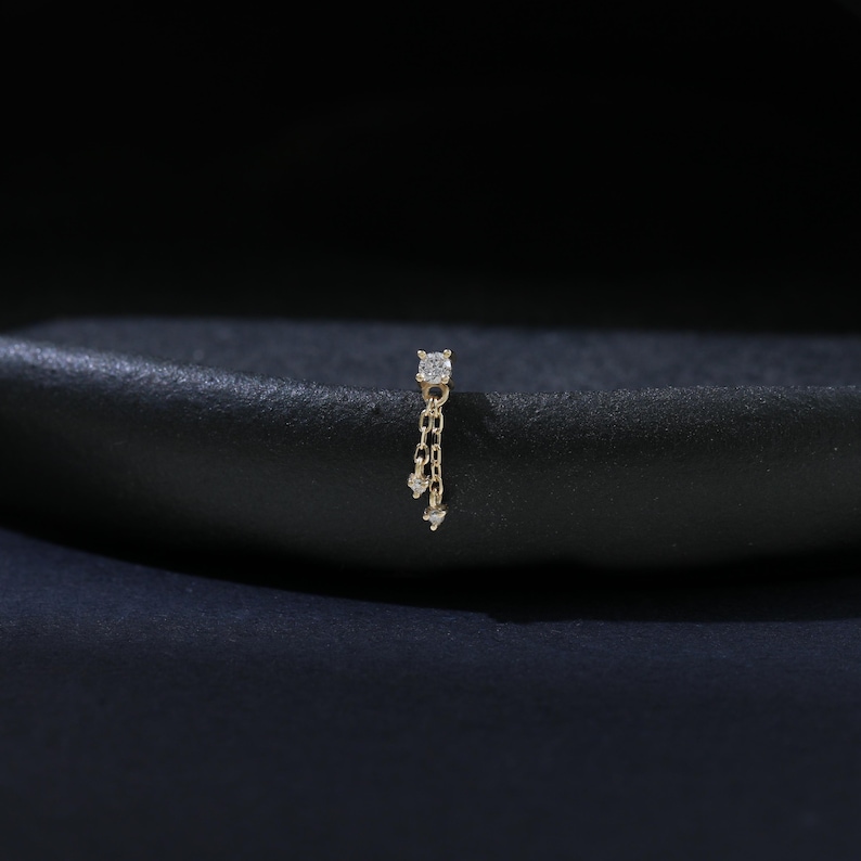 May include: A gold-colored nose stud featuring a square-cut, clear gemstone at the top, with a delicate chain suspending three smaller, clear gemstones. The jewelry is set against a dark background.
