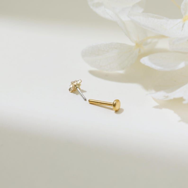 May include: Two gold nose rings, one with a small round ball and the other with a star-shaped design. The rings are on a white background.