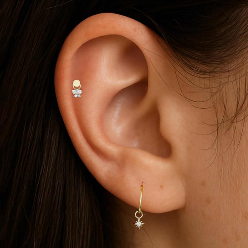Puede incluir: Primer plano de una oreja adornada con dos joyas. Un peque&ntilde;o pendiente de oro con dise&ntilde;o de flor en la parte superior de la oreja. Un pendiente de aro dorado con un dije de estrella cuelga del l&oacute;bulo de la oreja.