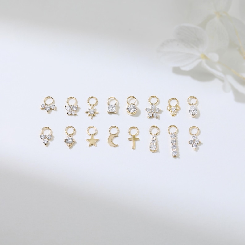 Puede incluir: Una colecci&oacute;n de colgantes dorados con varios dise&ntilde;os, incluyendo estrellas, flores y cruces. Cada colgante est&aacute; adornado con peque&ntilde;as gemas transparentes. Los colgantes est&aacute;n dispuestos sobre una superficie blanca.