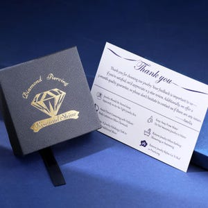 Puede incluir: Una caja de joyer&iacute;a gris oscuro con letras doradas que dicen "Diamond Piercing" y "Rise and Shine". Una tarjeta blanca con las palabras "Thank you" e instrucciones de cuidado est&aacute; al lado de la caja. El fondo es azul oscuro.