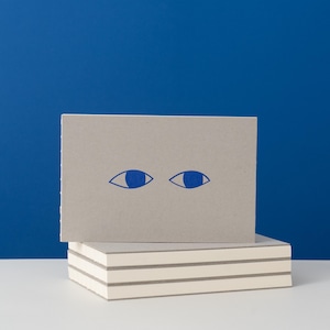 Könnte beinhalten: Ein Stapel Notizkarten mit einem blauen Augendesign auf einer grauen Karte. Die Karten sind auf einem Stapel leerer Karten gestapelt. Der Hintergrund ist einfarbig blau.