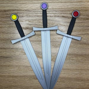 May include: Three toy swords with silver blades, black handles, and colorful pommels. The pommels are yellow, purple, and red. The swords are arranged in a fan shape on a wooden surface.