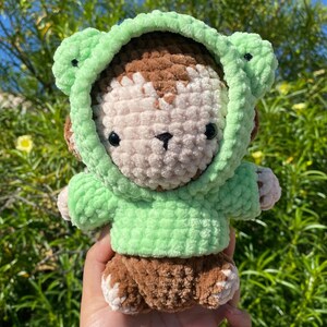 May include: A handmade crochet monkey doll wearing a light green frog hoodie. The monkey has brown and cream-colored fur, with black eyes and a small nose. The doll is held in a hand against a background of green foliage.