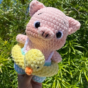 May include: A light pink crocheted pig plush toy featuring large black safety eyes and a snout. The pig is wearing a yellow crocheted duck-shaped swim ring around its midsection, which has a small orange beak and black eyes, over a pastel-colored outfit.