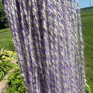 Puede incluir: Un conjunto de múltiples hebras de hilo colgando verticalmente. Cada hebra individual se compone de dos cabos distintos retorcidos, con una fibra púrpura vibrante entrelazada con una fibra suave de color gris claro y textura difusa.