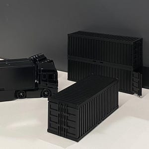 Puede incluir: Camión semirremolque y contenedores de envío de juguete negros. Los contenedores son rectangulares con líneas verticales y detalles en las esquinas. El camión es completamente negro.