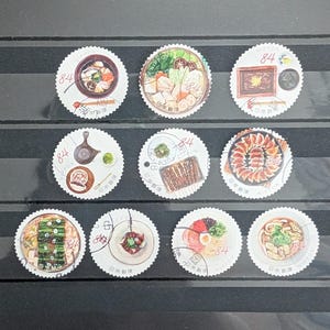 以下が含まれることがあります： 日本の食べ物をテーマにした10枚の切手のコレクション。各切手には、異なる料理のイラストが描かれており、「84」の数字と日本語のテキストが記載されています。切手は丸く、縁がスカラップになっており、暗い背景に配置されています。