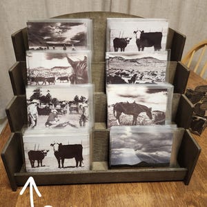 May include: A wooden display stand holding sets of black and white postcards. The postcards feature images of cows, horses, and landscapes. The word "Sets" is written on the front of the display.