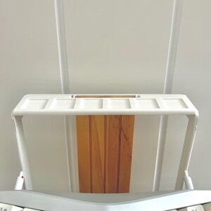 Puede incluir: Soporte de teclado de metal blanco con un estante de madera. El soporte tiene un marco de metal blanco y un estante de madera que está hecho de múltiples listones. El soporte está diseñado para sostener un teclado.