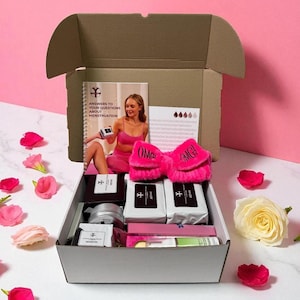 May include: An open cardboard box filled with various pink and white self-care products. Items include a booklet titled "Answers to Your Questions About Menstruation", fluffy pink slippers with "OMG" text, and several wrapped packages. Scattered flowers surround the box.