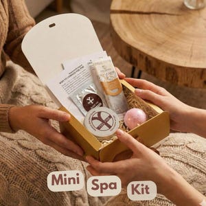 May include: A gold-colored mini spa kit box is held open, revealing spa products. The box contains a small tube of lotion, a bath bomb, a small round container, and other items. The text "Mini Spa Kit" is at the bottom.