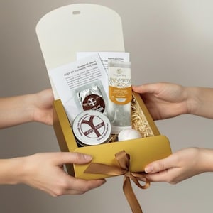 May include: A gold gift box tied with a brown ribbon, held by two sets of hands. Inside, a variety of items are visible, including a candle tin with "FEEL FINE" text, a tea tube, and a packet with text. The box is open, revealing its contents.