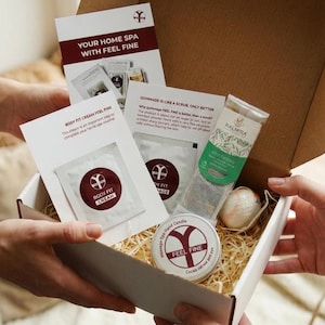 May include: A gift box filled with spa products, including a candle, tea, and body cream. The box contains printed cards with text that reads "YOUR HOME SPA WITH FEEL FINE" and "BODY FIT CREAM FEEL FINE". The candle is labeled "FEEL FINE".