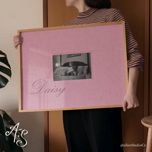 Puede incluir: Una foto enmarcada rosa con una foto en blanco y negro de un perro en una cama. El texto "Daisy" está escrito en cursiva debajo de la foto.