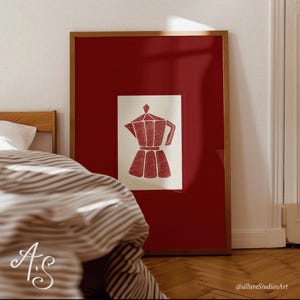 Puede incluir: Una impresión enmarcada con una ilustración de cafetera roja sobre un fondo crema, sobre un fondo rojo intenso. La obra de arte está encerrada en un marco de madera. La imagen se muestra en un dormitorio.