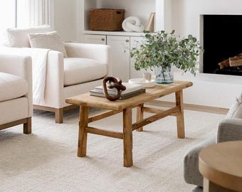 May include: A rectangular wooden coffee table with four legs and a horizontal support beam, featuring a natural wood grain finish. On the table sits a stack of books, a dark brown decorative wooden knot, and a glass vase filled with green eucalyptus.
