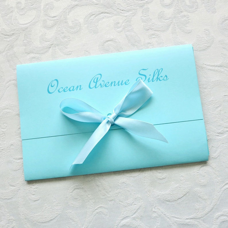 Gift packaging. Aqua blue folded envelope with aqua blue bow. Ocean Avenue Silks in script font above bow.