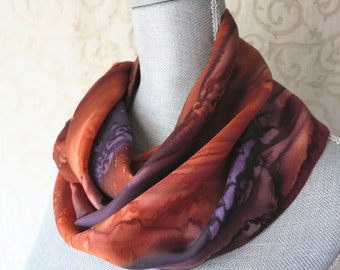 May include: A silk infinity scarf in shades of rust, purple, and brown, draped on a mannequin. The scarf features a marbled pattern with flowing color transitions. The fabric appears soft and lightweight, suitable for various seasons.