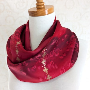 May include: A red infinity scarf with a floral pattern and gold accents. The scarf is displayed on a mannequin and has a soft, flowing appearance. The design features a gradient of red hues, creating a visually appealing accessory.