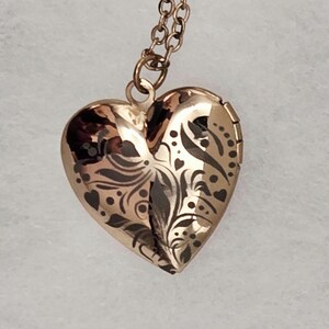 May include: A gold-colored heart-shaped locket pendant with a decorative floral and heart pattern. The locket hangs from a delicate chain. The locket is closed and appears to be made of metal.