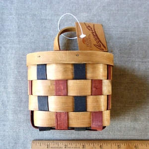 May include: A small, wooden wall basket with a black and red woven pattern. The basket has a handle and a tag that reads "Home Creations Accents".