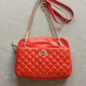 Pu&ograve; includere: Borsa a tracolla trapuntata rossa con tracolla a catena dorata e dettagli borchiati. Presenta un manico superiore e una chiusura decorativa. La borsa &egrave; rettangolare e ha una chiusura a zip.