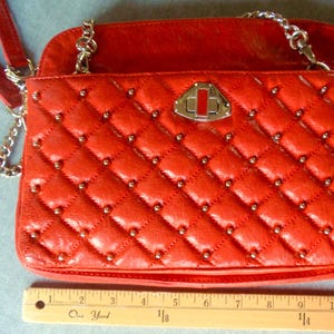 Pu&ograve; includere: Borsa a tracolla rossa trapuntata con catene argentate e borchie. La borsa ha una forma rettangolare con chiusura a patta e chiusura argentata. La borsa &egrave; larga circa 25 cm.