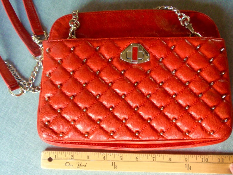 Pu&ograve; includere: Borsa a tracolla rossa trapuntata con catene argentate e borchie. La borsa ha una forma rettangolare con chiusura a patta e chiusura argentata. La borsa &egrave; larga circa 25 cm.