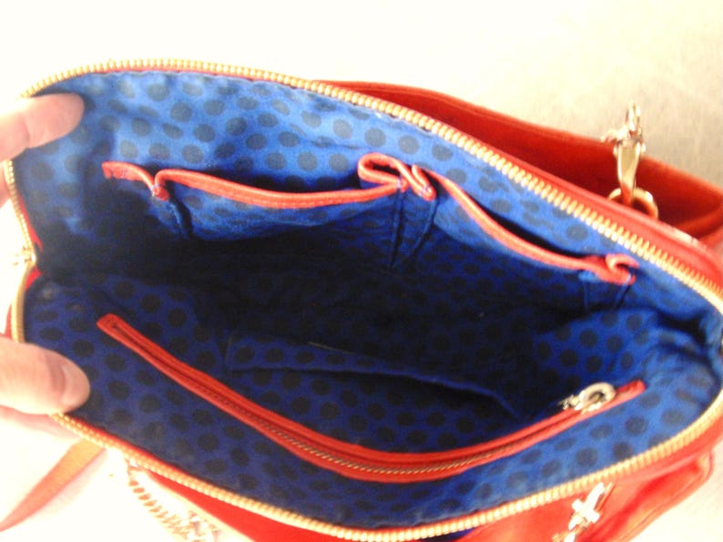 Pu&ograve; includere: Vista interna di una borsa rossa con cerniera dorata. L'interno &egrave; blu con un motivo a pois neri e ha diverse tasche. La borsa ha una tracolla a catena dorata.