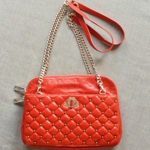 Pu&ograve; includere: Una borsa rossa trapuntata con borchie color oro e una tracolla a catena dorata. La borsa ha una forma rettangolare con chiusura a patta e un piccolo manico in pelle rossa. La borsa &egrave; su una superficie grigia.