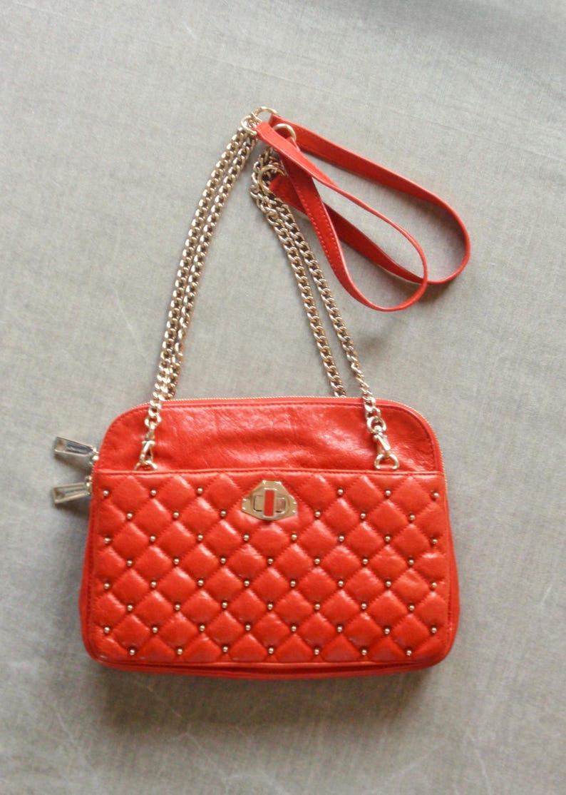 Pu&ograve; includere: Una borsa rossa trapuntata con borchie color oro e una tracolla a catena dorata. La borsa ha una forma rettangolare con chiusura a patta e un piccolo manico in pelle rossa. La borsa &egrave; su una superficie grigia.