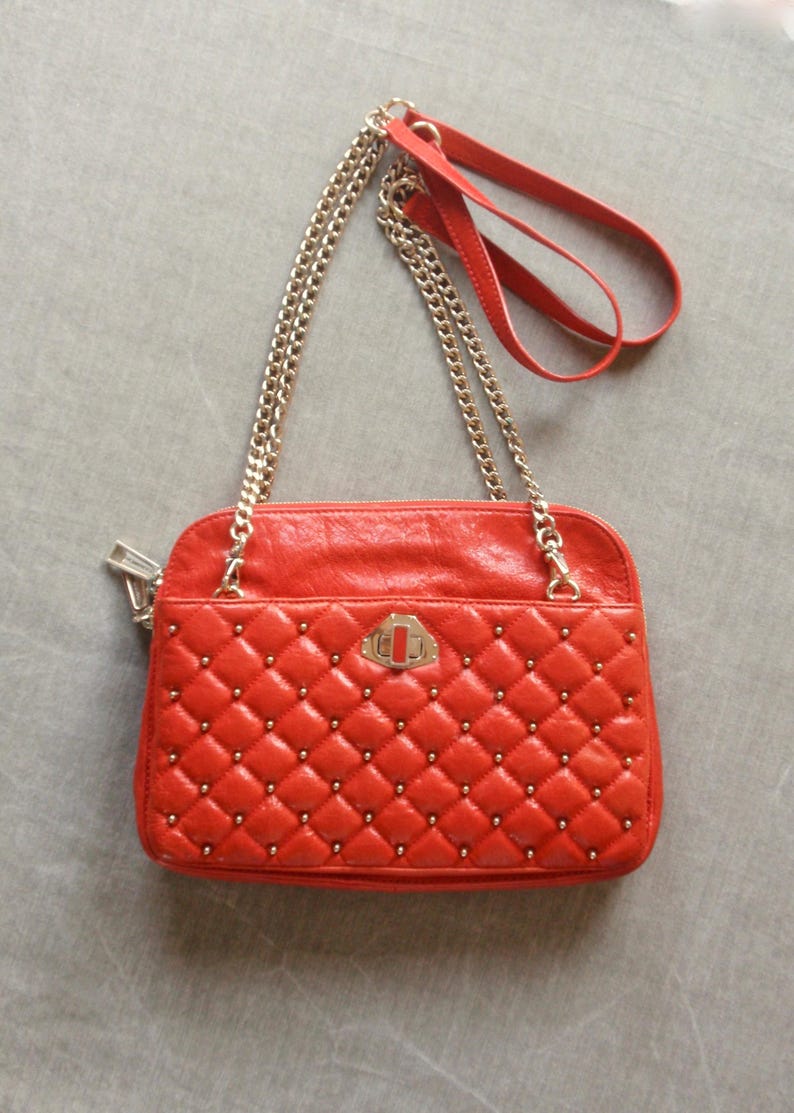 Pu&ograve; includere: Borsa a tracolla trapuntata rossa con tracolla a catena dorata e dettagli borchiati. Presenta un manico superiore e una chiusura decorativa. La borsa &egrave; rettangolare e ha una chiusura a zip.